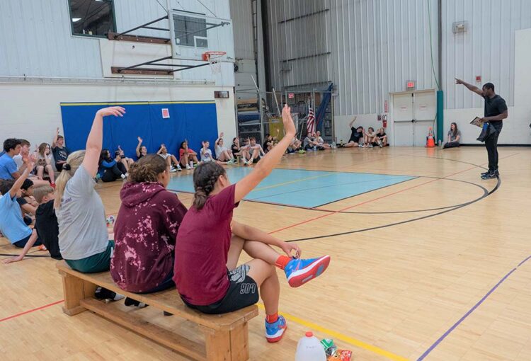 Man speaking to half-circle of basketball campers with hands raised