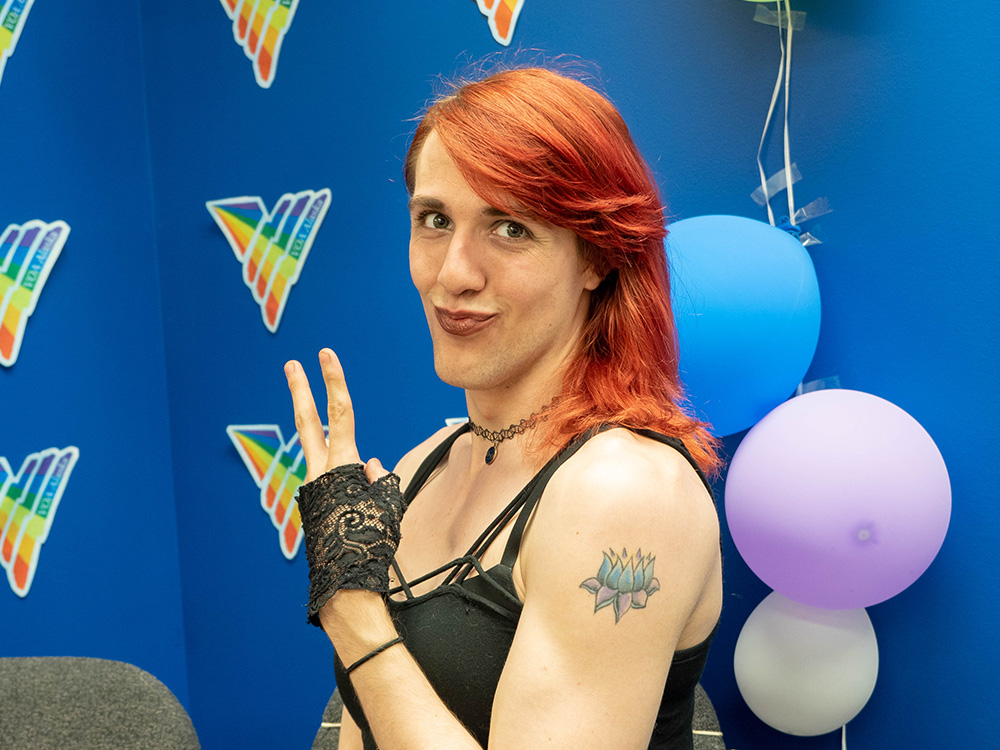Person with long, bright red hair giving a peace sign in front of a wall decorated for Pride Month