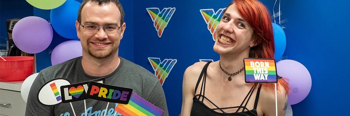 Two people smiling in front of a Pride Month decorated wall