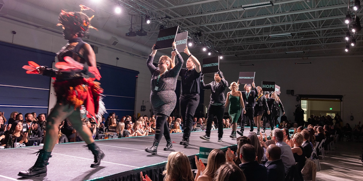 Group of youth and adults on stage at a fashion show holding signs with core values following a model dressed as a phoenix