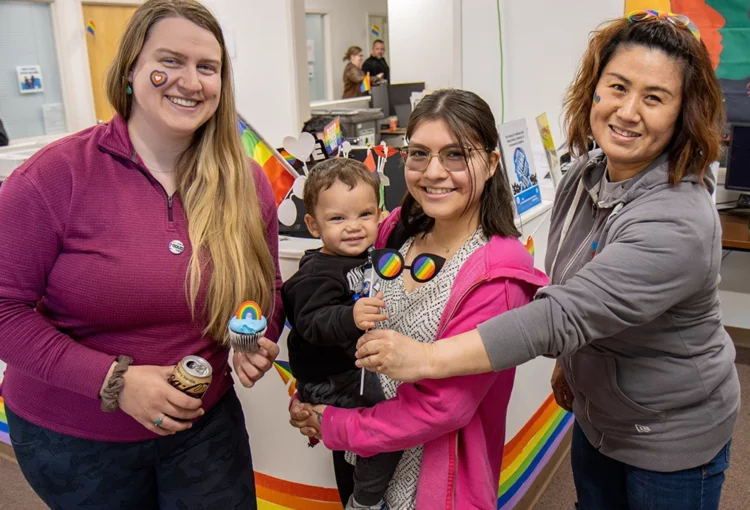 Three people pose with pride month props, the one in the middle is holding a young child with a large grin.