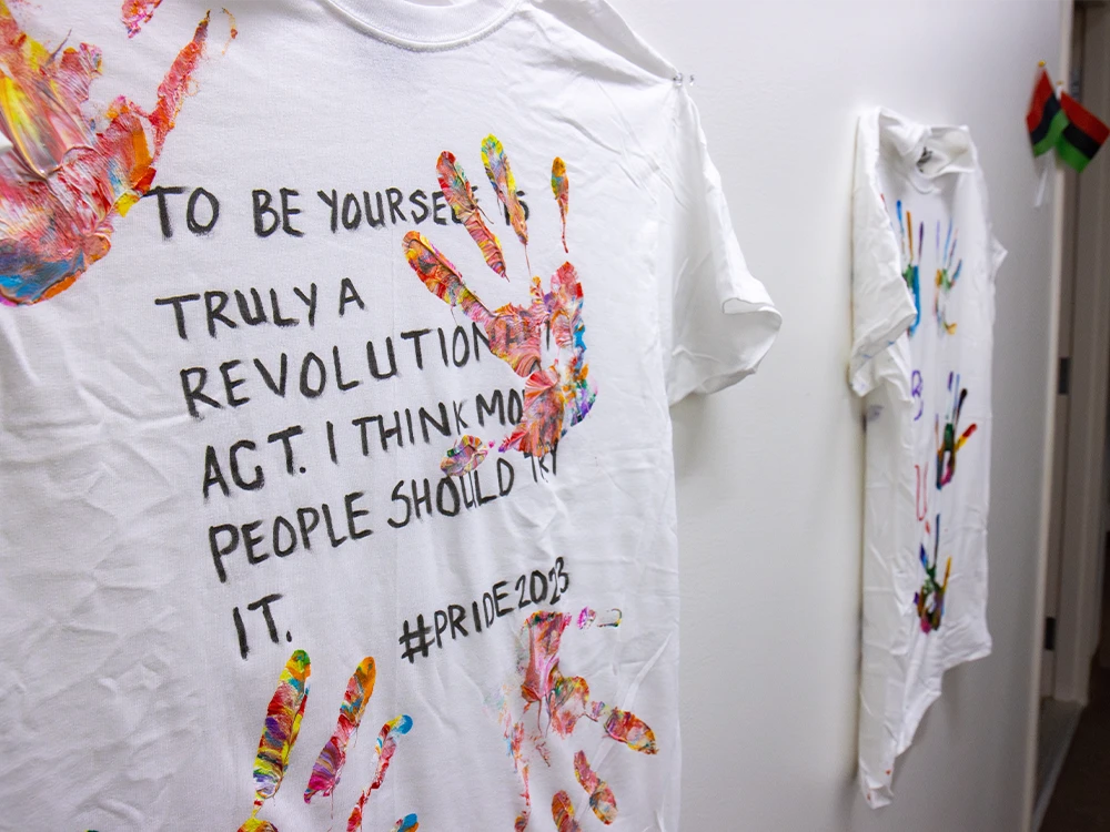 A shirt hangs on a wall, with colorful, painted handprints and the words: To be yourself is truly a revolutionary act. I think more people should try it.