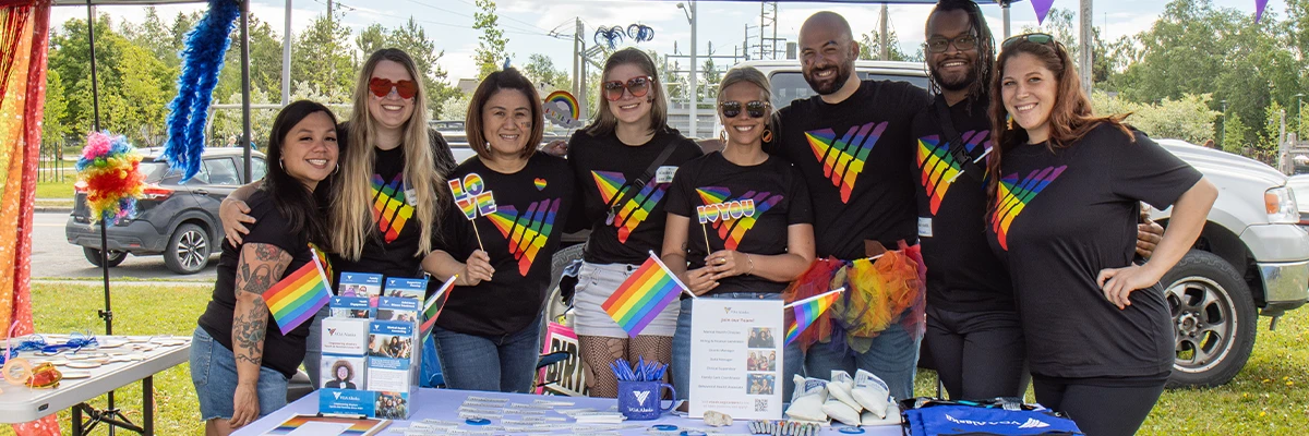 Group of 8 people at a table decorated for Pride Month. They are each wearing a black t-shirt with a rainbow colored V from the VOA logo.