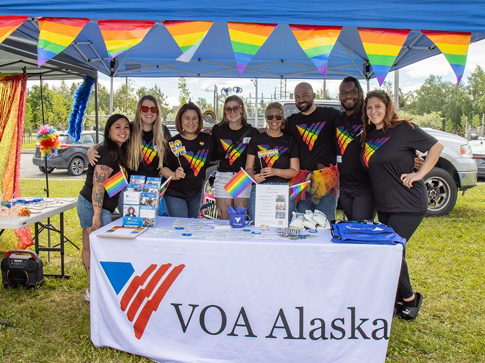8 VOA staff, wearing rainbow colored V-logos on black t-shirts, pose behind the VOA table
