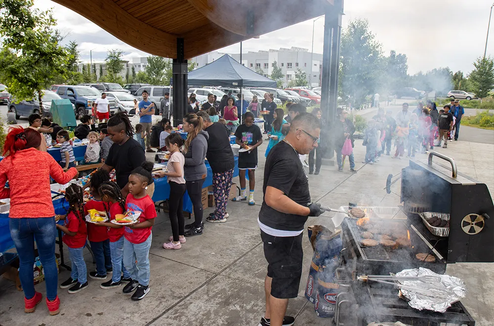 In the foreground, a person stands at a smoky grill flipping burgers. In the background, a line of people prepare plates while more people wait behind them.
