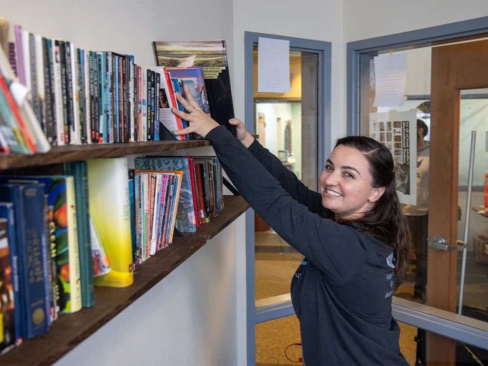 Person smiling at camera while adding books to a shelf.
