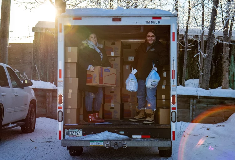 Two people inside a U-haul moving truck in a snowy parking lot. The person on left holds a box, the person on the right holds up two frozen turkeys. Behind them are dozens of more boxes.