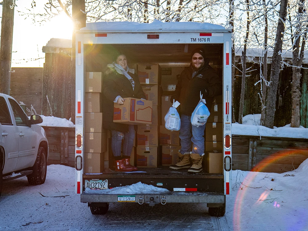 Two people inside a U-haul moving truck in a snowy parking lot. The person on left holds a box, the person on the right holds up two frozen turkeys. Behind them are dozens of more boxes.