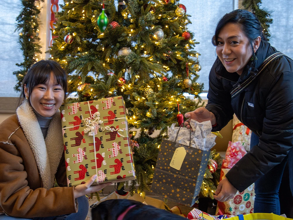 Two people posing with gifts in front of a Christmas tree.