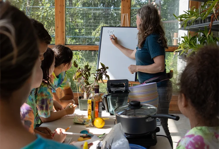 In a sun-filled room, a group of kids watch an instructor writing on a white board.