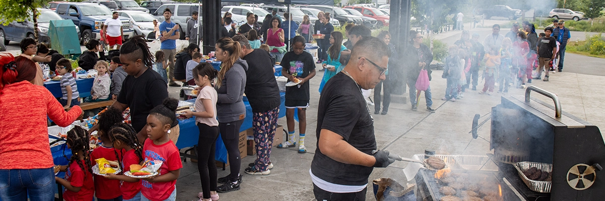A community barbecue with a line around the food table and someone at a smoking barbecue.