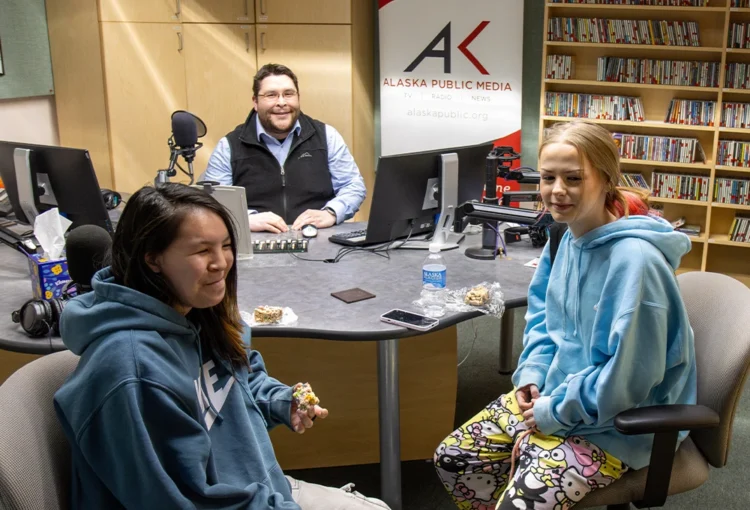 An adult with two youth, sitting in recording studio, with the Alaska Public Media logo behind them.