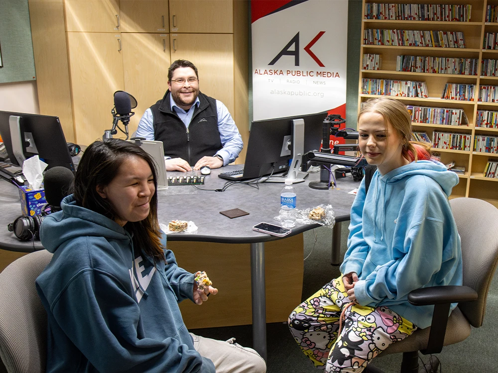 An adult with two youth, sitting in recording studio, with the Alaska Public Media logo behind them.