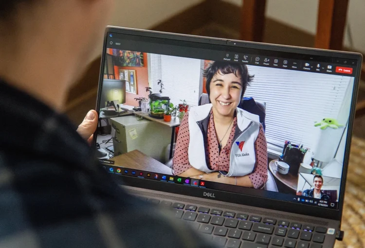 Over-the shoulder image depicting a telehealth counseling session with a person looking at a laptop screen displaying a video call with someone wearing a VOA Alaska vest