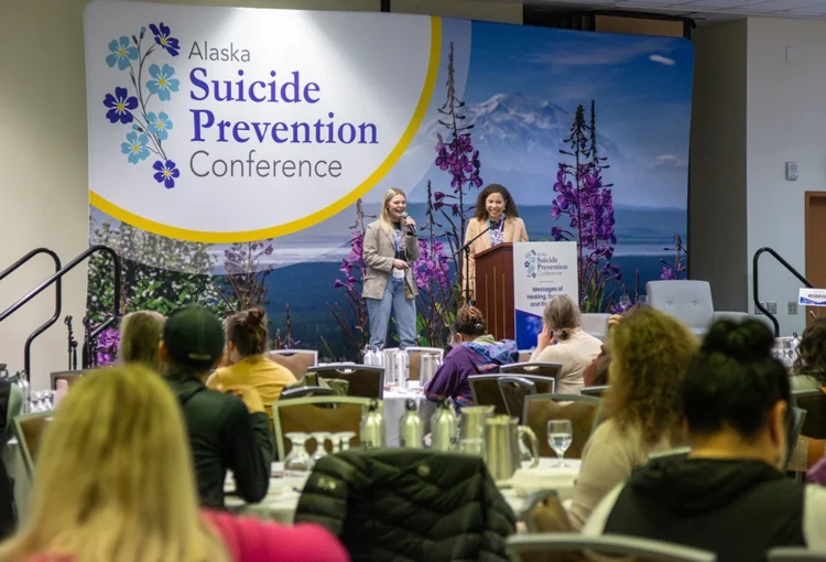 Two speakers on a stage with a large sign behind them reading "Alaska Suicide Prevention Conference."