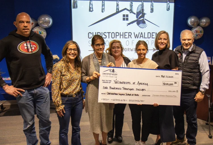 A group of people stand in front of a projected logo for the Christopher Walden School of Hope while holding a large check for the amount of $100,000.