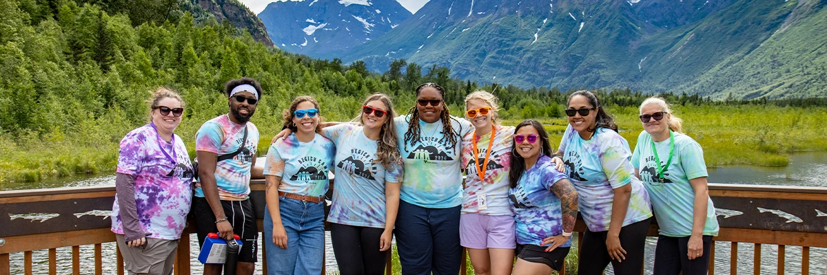 A group of staff dresses in tie-dye shirts and bright sunglasses on a boardwalk in front of mountains.