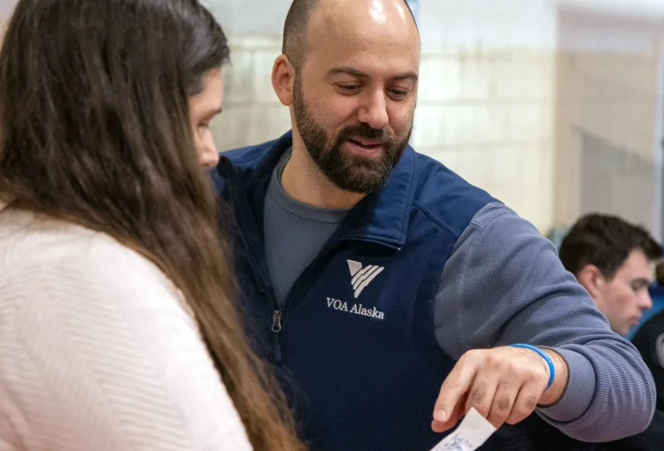 A person wearing a blue VOA Alaska-branded vest puts to a piece of paper being held by an unseen person.