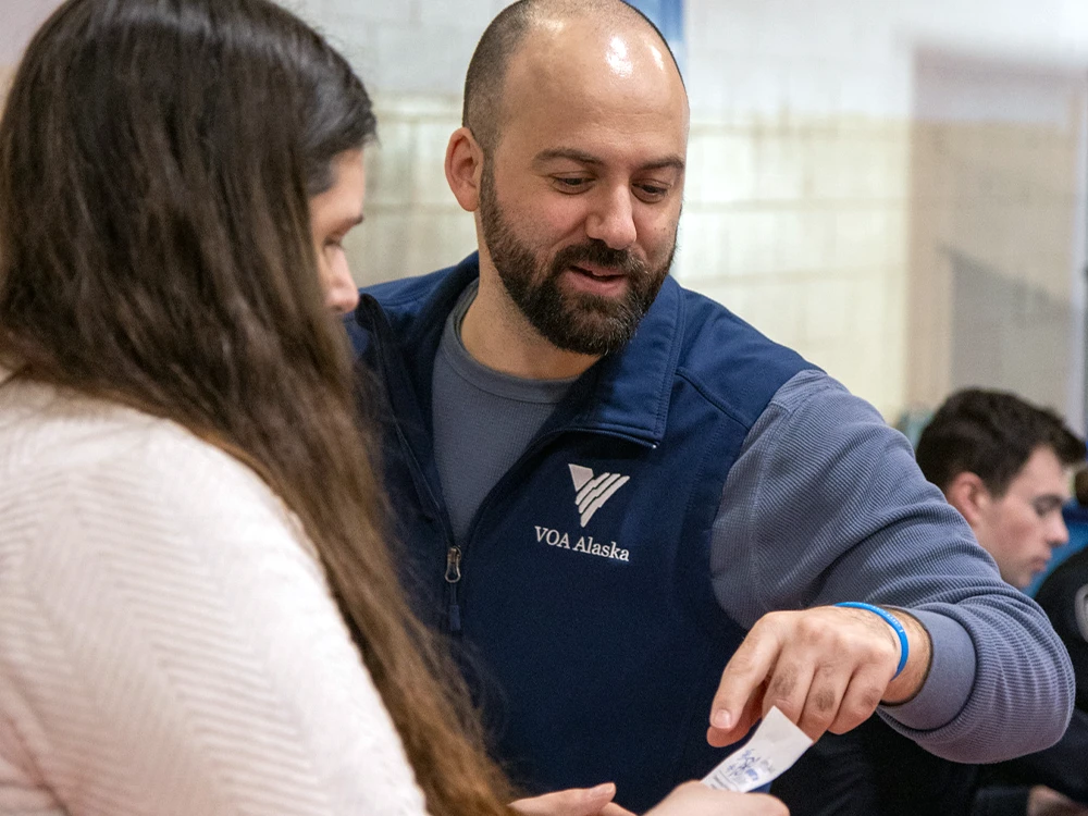 A person wearing a blue VOA Alaska-branded vest puts to a piece of paper being held by an unseen person.